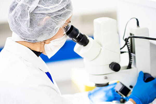 Mujer con lentes cofia, cubrebocas y bata blanca, analizando muestra en microscopio dentro de Laboratorio Químico de Tecnoalimentos, para el cuidado de la calidad e inocuidad de la materia prima producida por Tecnoalimentos en Querétaro