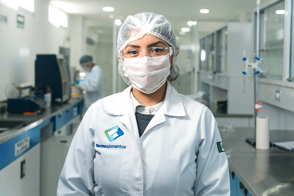 Mujer con lentes, cofia, cubrebocas y bata, dentro de Laboratorio Químico de Tecnoalimentos, viendo al frente, después de realizar análisis fisicoquímico y de biología molecular en México